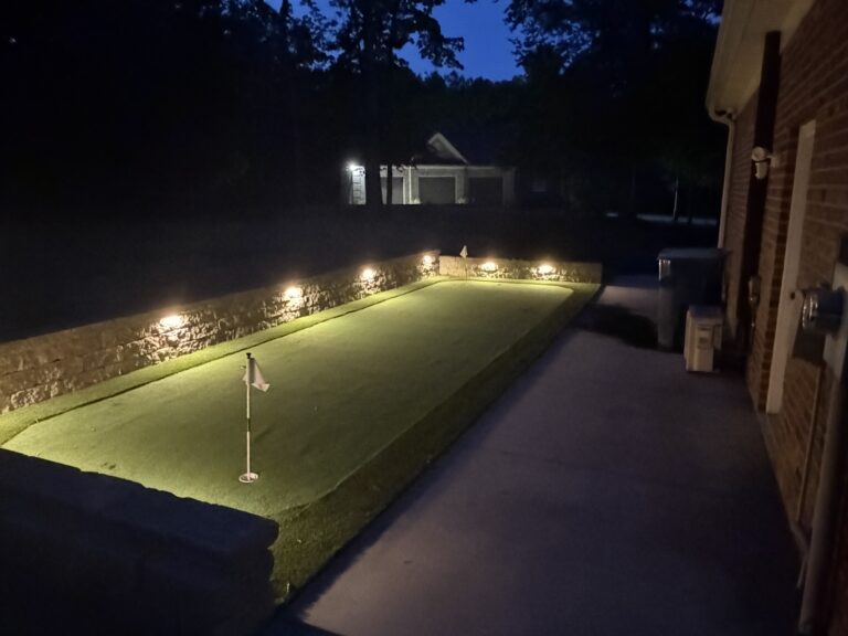 Landscape lighting fixtures mounted on a stone retaining wall illuminating a synthetic backyard putting green at night