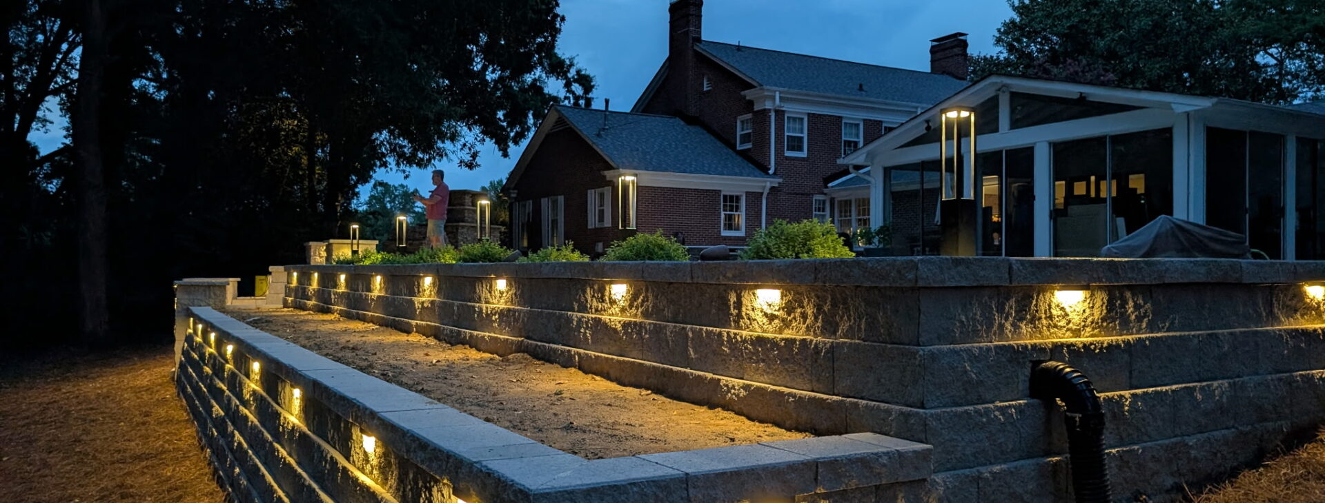 Landscape lighting fixtures spaced along a multi-tier stone retaining wall in front of a brick home at dusk