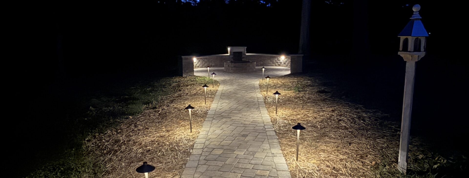 Row of path lights along a stone paver walkway leading to a lit stone fire pit seating area