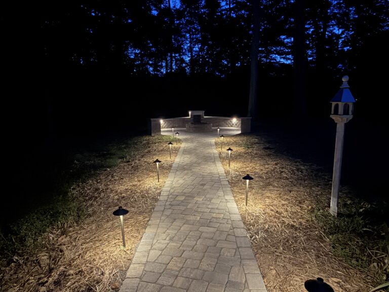 Row of path lights along a stone paver walkway leading to a lit stone fire pit seating area