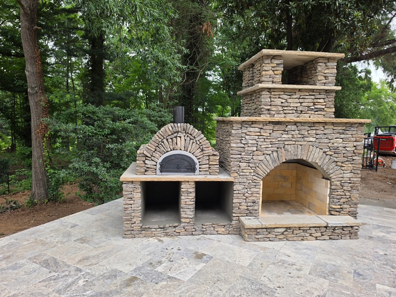 Custom stacked stone outdoor fireplace with attached pizza oven on travertine patio — Garrison Landscaping