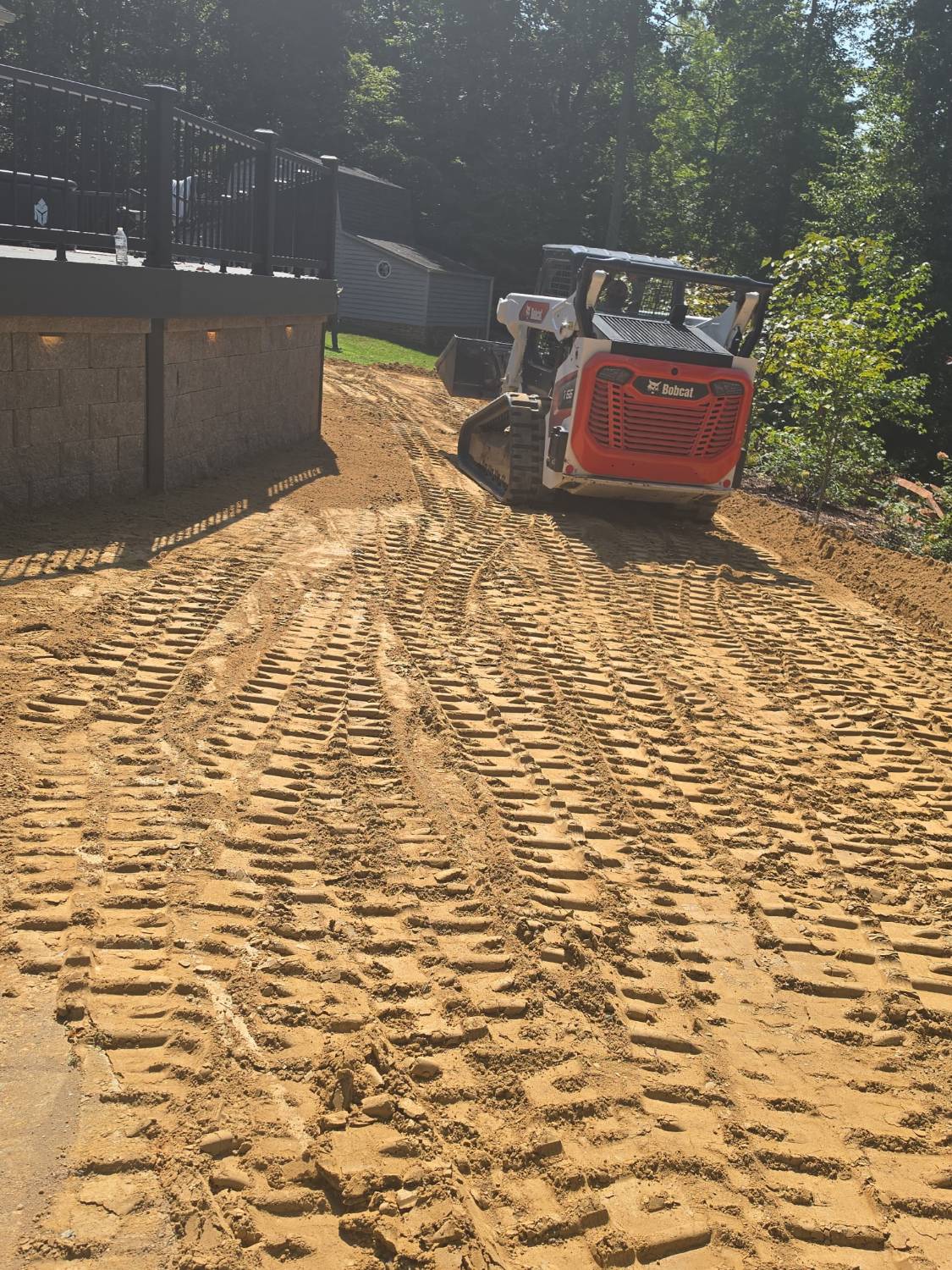 Bobcat track loader grading a yard beside a deck and retaining wall