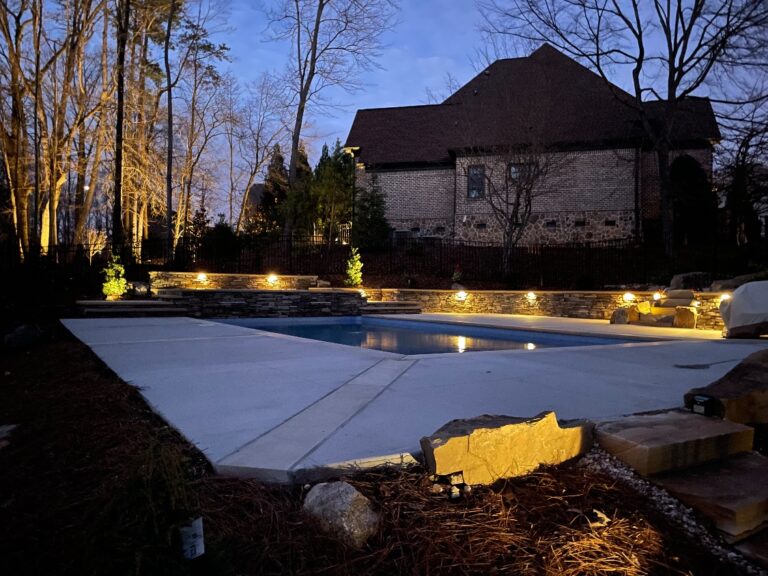Concrete pool deck with stacked-stone retaining walls and wall cap lighting at dusk