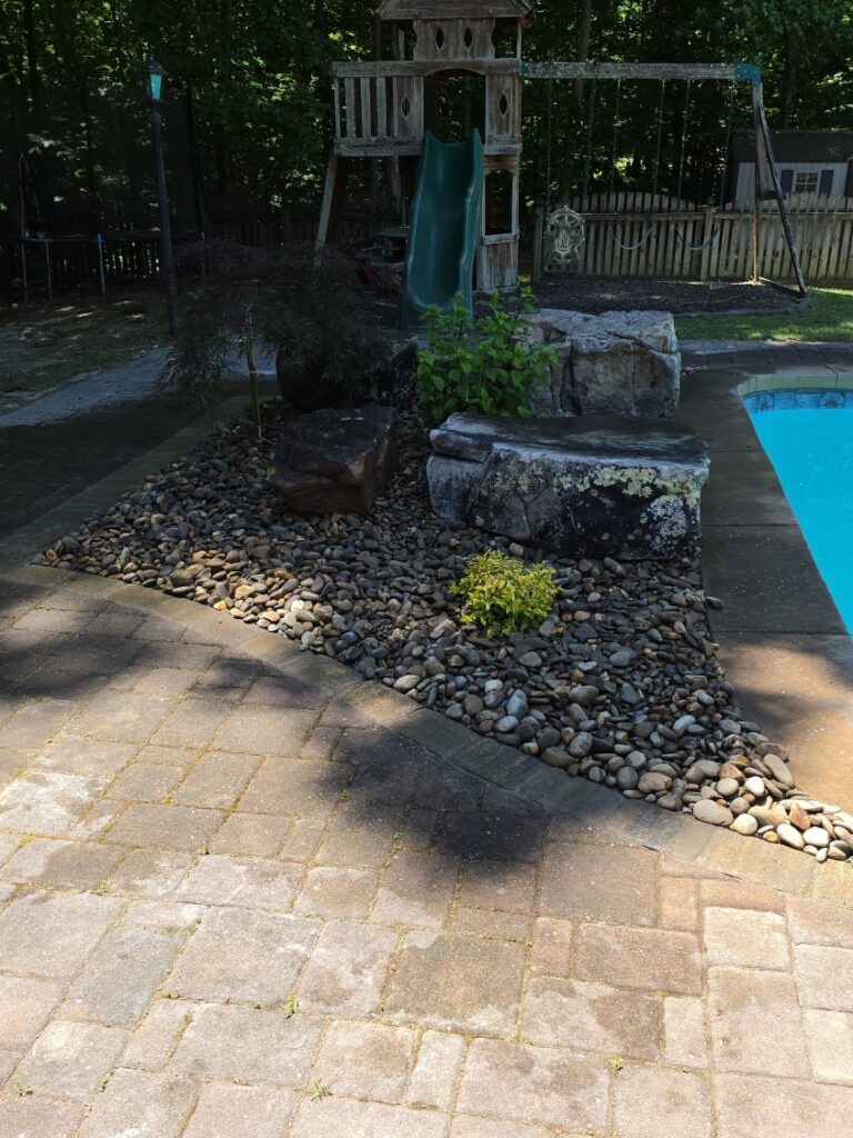 Poolside planting bed of river rock with natural boulder seating beside a paver pool deck