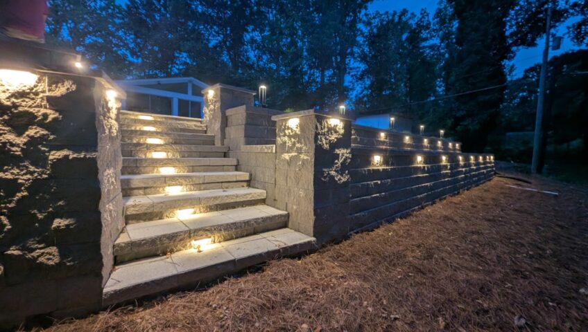 Segmental block retaining wall and stone steps with built-in step and wall cap lighting at dusk