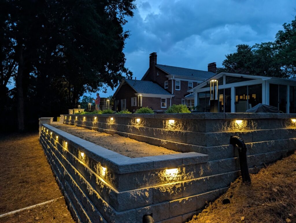 Retaining Walls 101: When You Need One and How They’re Built to Last 5 Two-tier block retaining wall with under-cap lighting below a raised backyard patio at dusk