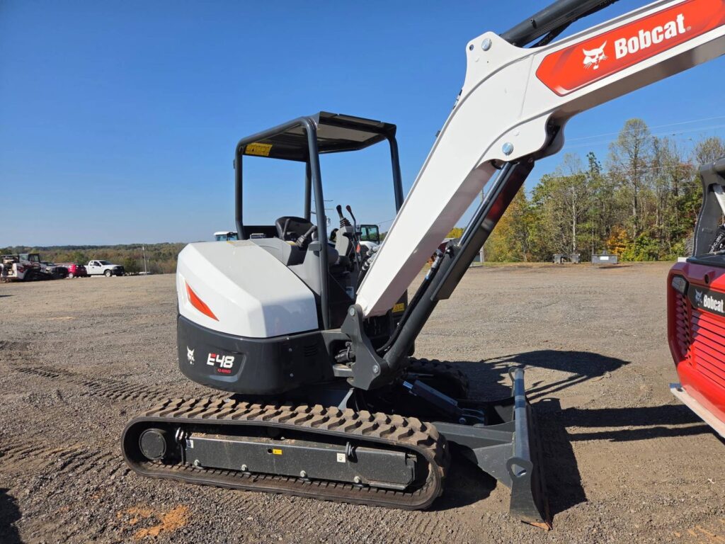 Bobcat E48 mini excavator with rubber tracks, an open canopy and a front blade, parked on a gravel lot