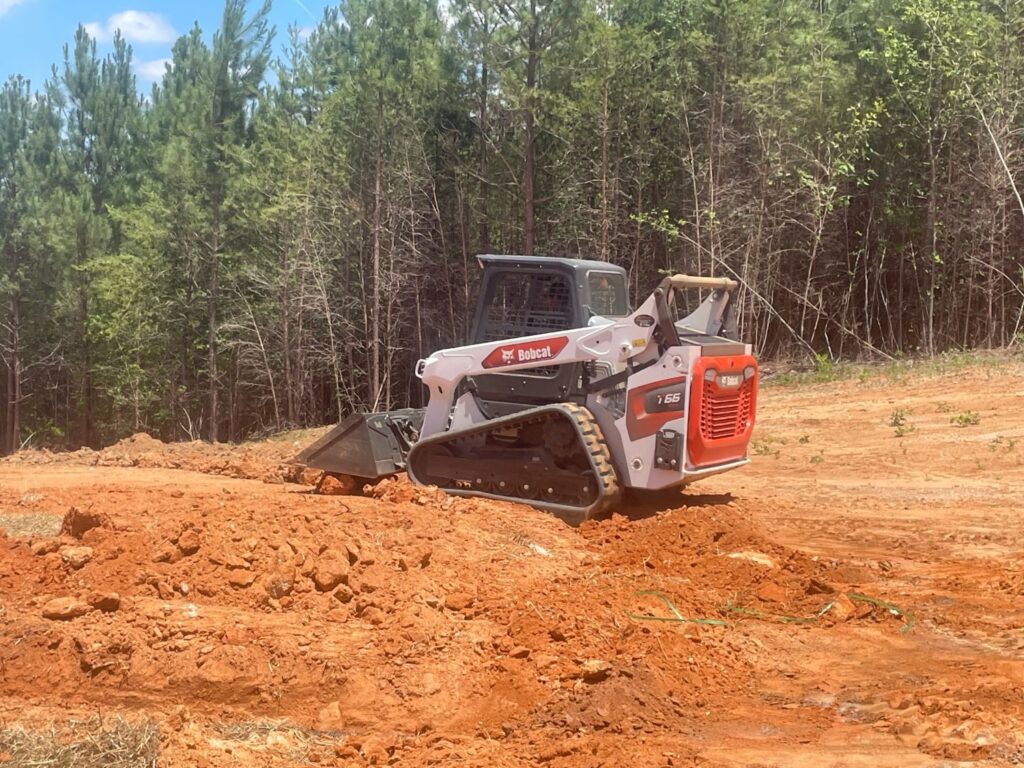 Bobcat T66 compact track loader grading red clay with its bucket on a cleared lot beside pine woods