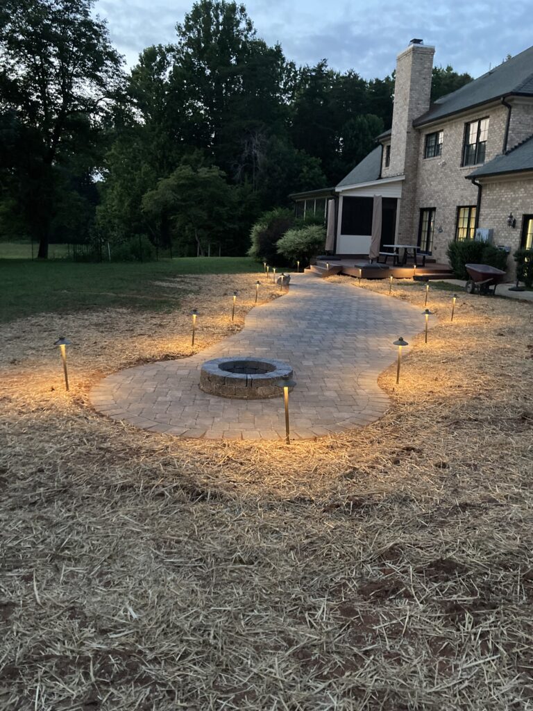 Curved paver patio with stone fire pit and low-voltage path lighting at dusk in Colfax, NC by Garrison's Landscaping
