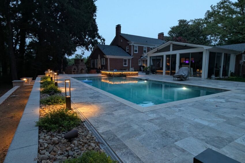 Travertine paver pool patio with a stacked-stone wall and landscape lighting at dusk, built by Garrison's Landscaping