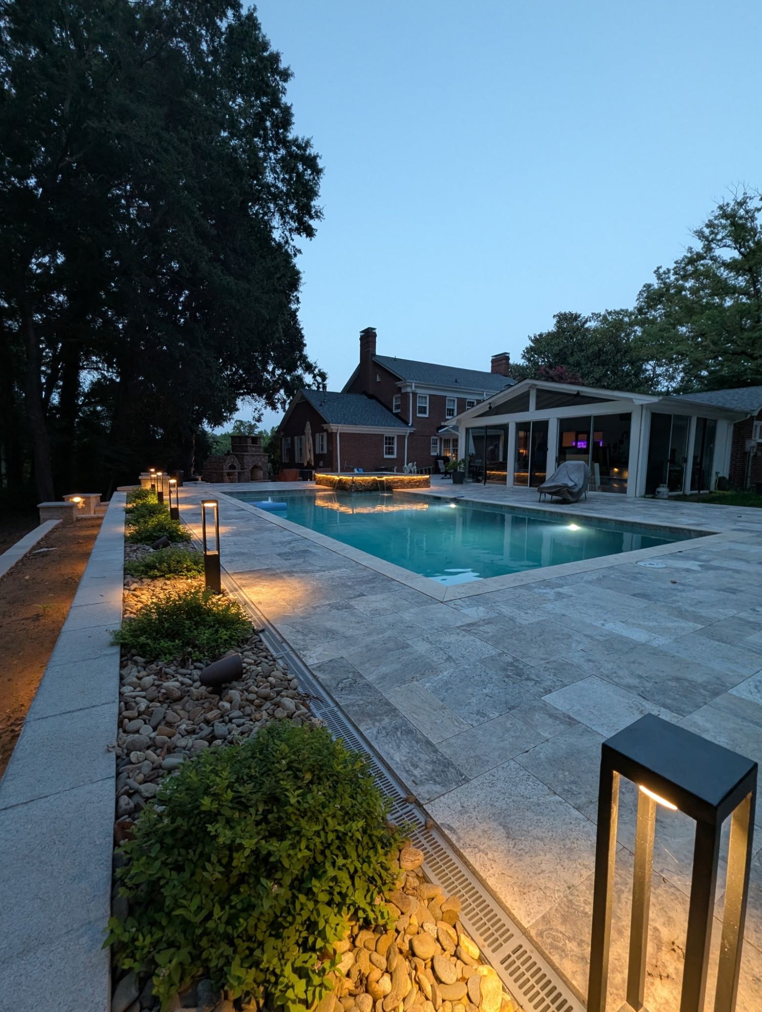 Travertine paver pool patio with a stacked-stone wall and landscape lighting at dusk, built by Garrison's Landscaping